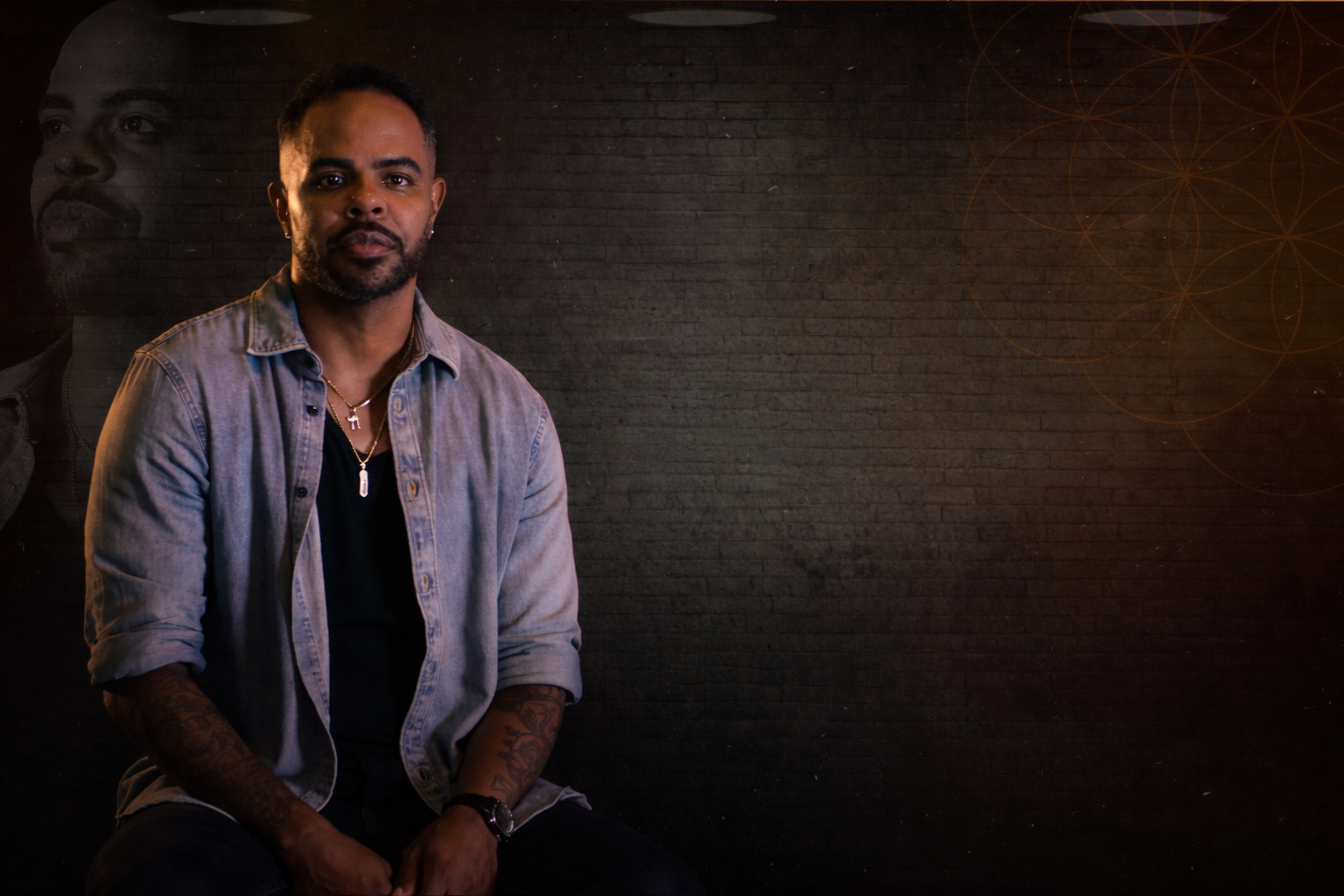 Sean Clayton seated against a brick wall in a denim shirt, crystal pendant visible. The host of the room.